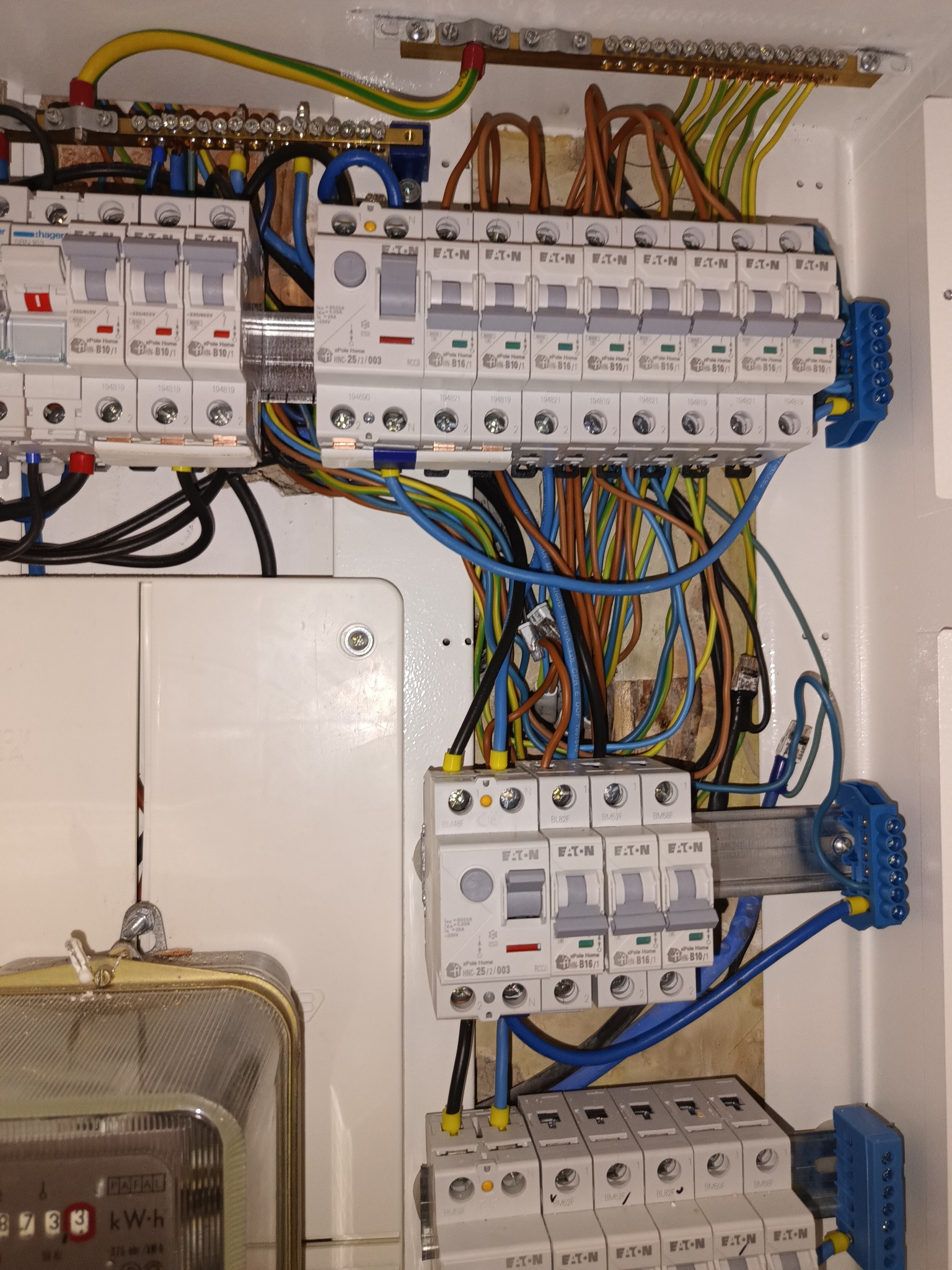 Rozdzielnia elektryczna w białej obudowie z widocznymi bezpiecznikami, przewodami w różnych kolorach i licznikiem energii elektrycznej po lewej stronie.