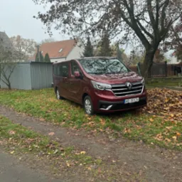 Czerwony Renault Trafic na tle jesiennego krajobrazu z opadłymi liśćmi. Auto zaparkowane na trawie, widoczny fragment domu i drzewa.