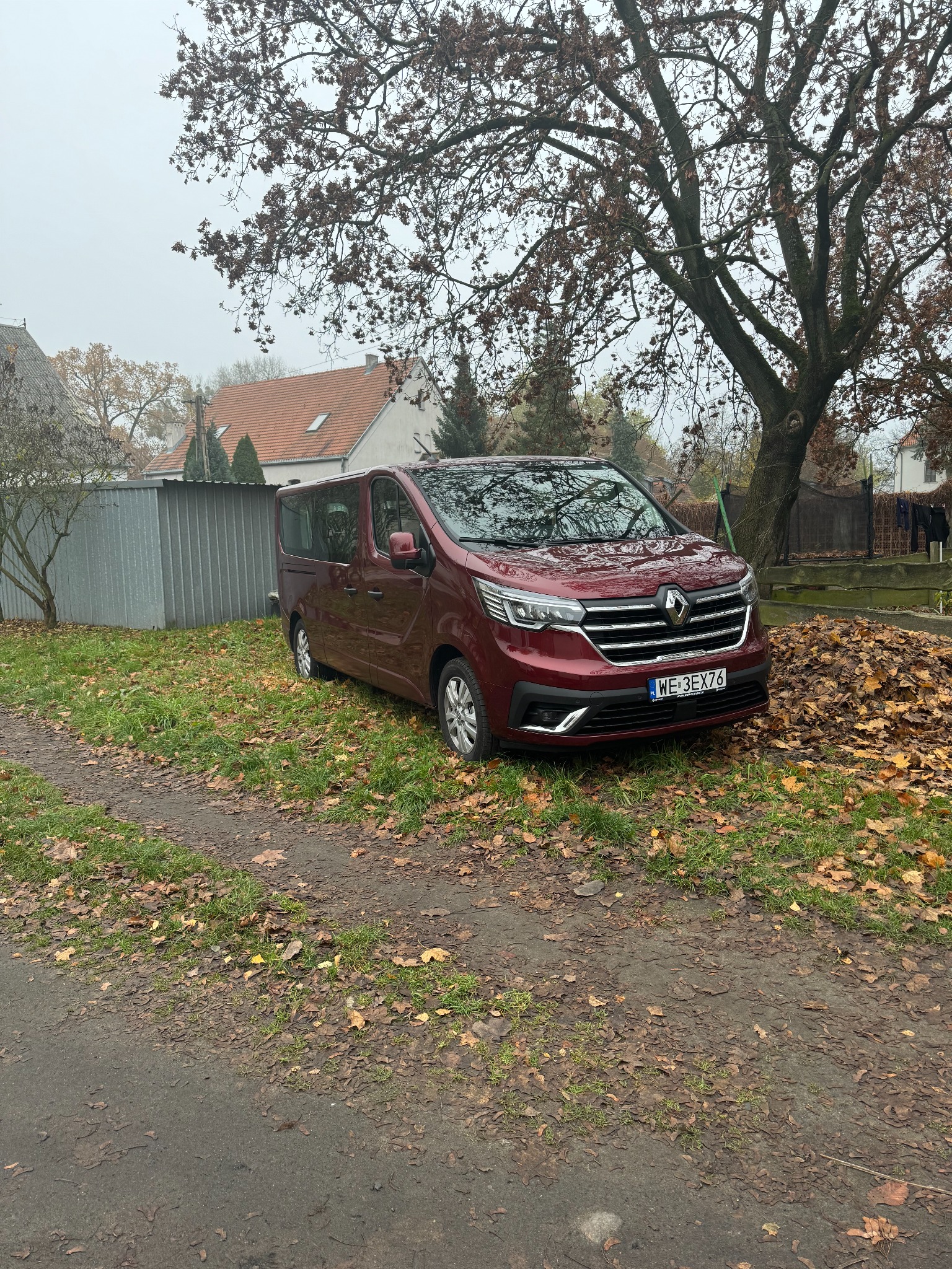 Czerwony Renault Trafic na tle jesiennego krajobrazu z opadłymi liśćmi. Auto zaparkowane na trawie, widoczny fragment domu i drzewa.