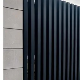 Nowoczesne ogrodzenie z pionowych, grafitowych metalowych profili, przylegające do betonowego murku z szarych bloczków. Minimalistyczny design.