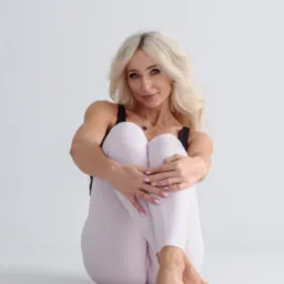Uśmiechnięta blondynka w sportowym stroju siedzi na podłodze, obejmując kolana. Jasne tło, naturalne światło, widoczny manicure. Prezentacja elastyczności i dobrej kondycji.