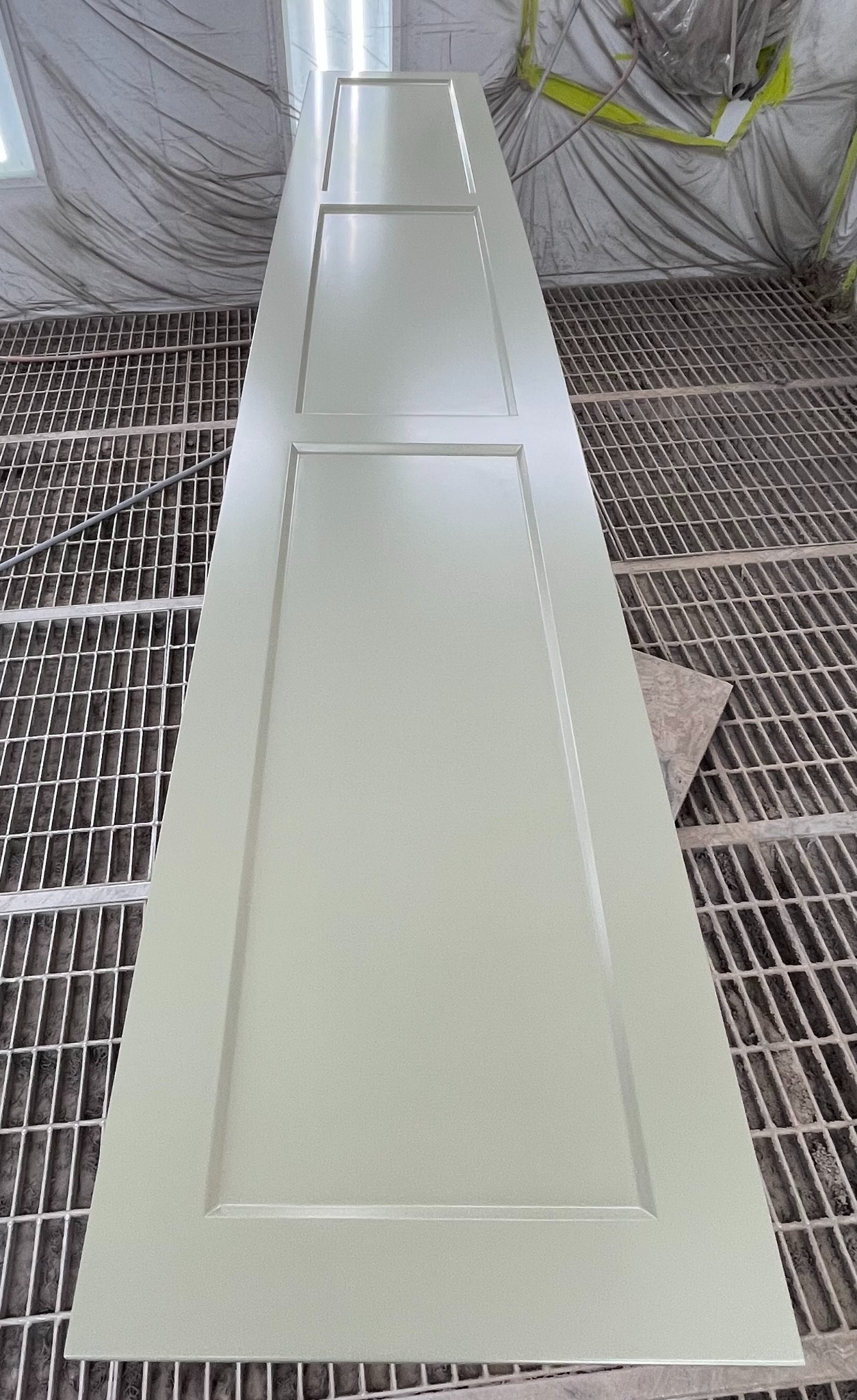 Świeżo pomalowane, kremowe drzwi z trzema panelami, leżące na metalowej kratownicy w warsztacie lakierniczym, ukazujące gładką powierzchnię i precyzyjne wykończenie.