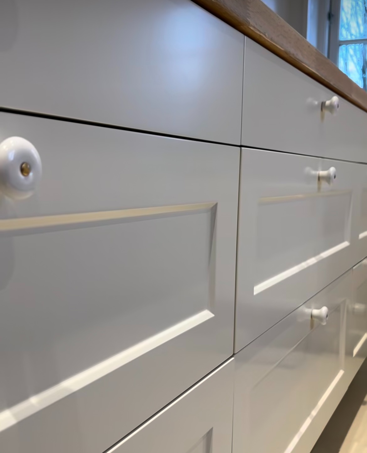 Białe, matowe fronty szuflad kuchennych z frezowanymi ramkami i okrągłymi, porcelanowymi uchwytami. Blat z jasnego drewna. Ujęcie z dołu, perspektywa zbieżna.