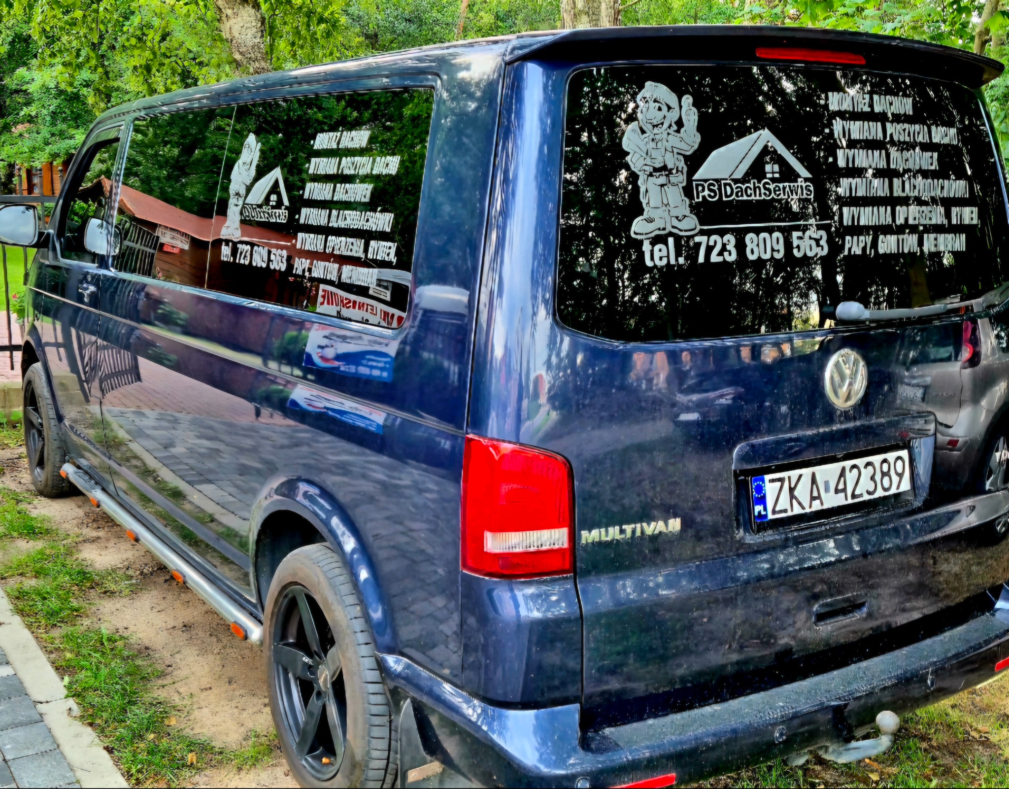 Granatowy bus firmy dekarskiej z reklamą usług na szybach: montaż, wymiana pokryć dachowych, rynien. Widoczny numer telefonu i logo firmy PS Dach Serwis.