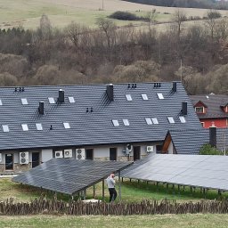 M-tech Mateusz Kasperkiewicz - Instalacja paneli słonecznych na stelażach przed budynkiem z dachem pokrytym dachówką, w tle pagórkowaty krajobraz. Osoba przy panelach.