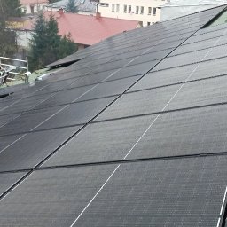 M-tech Mateusz Kasperkiewicz - Montaż paneli fotowoltaicznych na zielonym dachu. Widoczny pracownik na rusztowaniu i zabudowa miejska w tle. Pochmurny dzień.