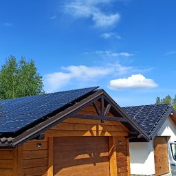 M-tech Mateusz Kasperkiewicz - Panele słoneczne na dachu drewnianego garażu i białego domu w słoneczny dzień. Na pierwszym planie samochód dostawczy, w tle zieleń drzew i błękitne niebo.