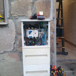 M-tech Mateusz Kasperkiewicz - Otwarta skrzynka elektryczna na kamiennej ścianie budynku, widoczne kable, bezpieczniki i narzędzia elektryczne. Obok drabina i torba z narzędziami.