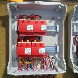 M-tech Mateusz Kasperkiewicz - Rozdzielnica z czerwonymi ogranicznikami przepięć i okablowaniem w kolorach czarnym, czerwonym i żółtym. Widoczne elementy montażowe i śruby.