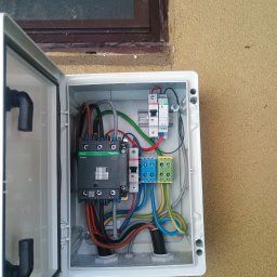 M-tech Mateusz Kasperkiewicz - Otwarta, biała skrzynka elektryczna z widocznymi bezpiecznikami, stycznikiem Schneider Electric i kolorowymi przewodami. Skrzynka zamontowana na elewacji budynku obok okna.