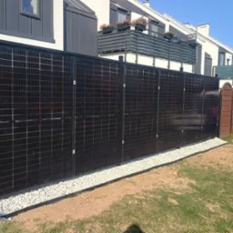 Panele fotowoltaiczne zamontowane jako ogrodzenie posesji. Widoczne czarne ramy, jasne kamienie u podstawy i fragmenty budynków w tle, w słoneczny dzień.
