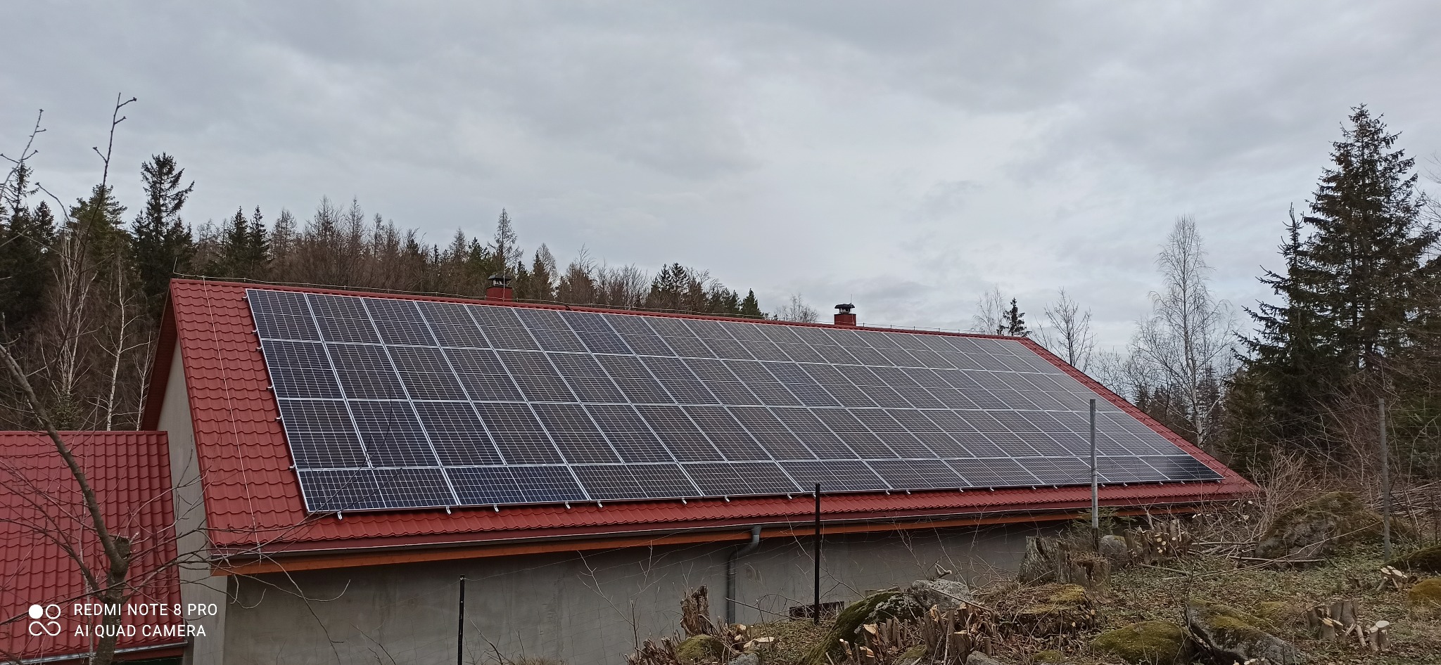 Dom z czerwoną dachówką i panelami słonecznymi na dachu, otoczony drzewami. Widok na dużą instalację fotowoltaiczną w pochmurny dzień.