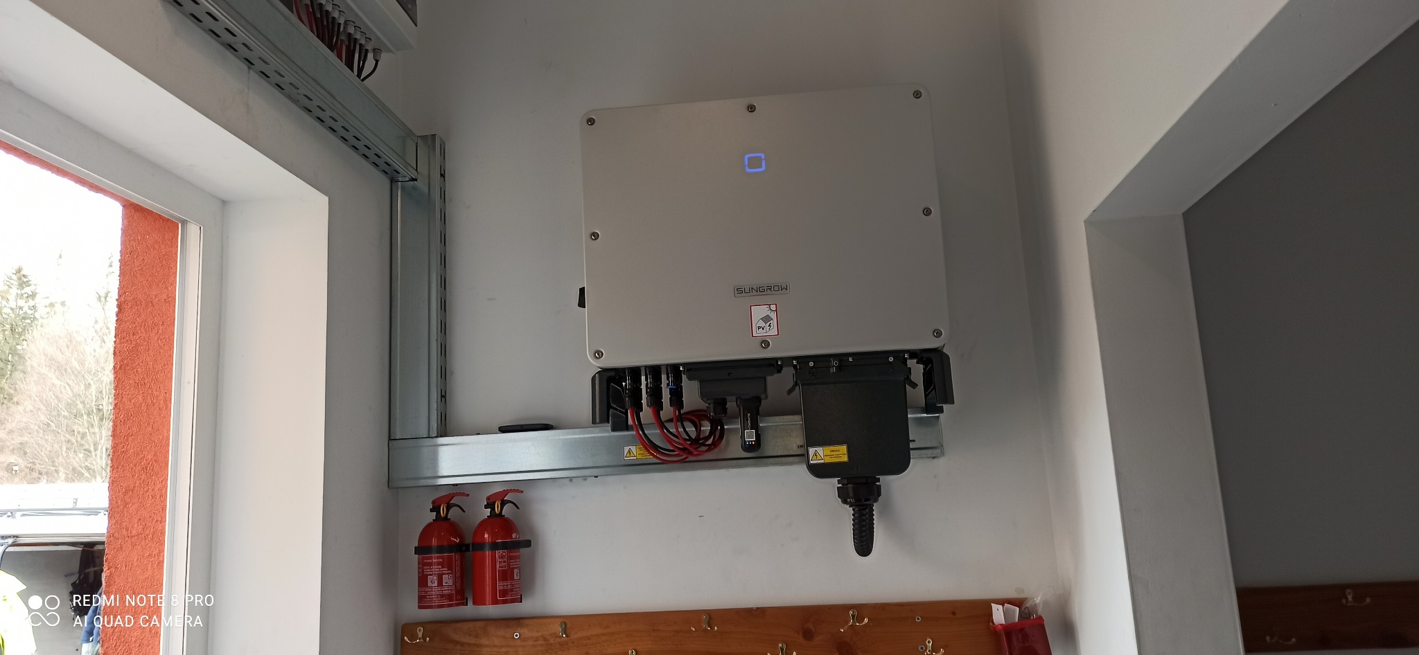 Zamontowany inwerter fotowoltaiczny Sungrow na ścianie, widoczne przewody, zabezpieczenia i fragment okna. Dwa czerwone gaśnice pod inwerterem.