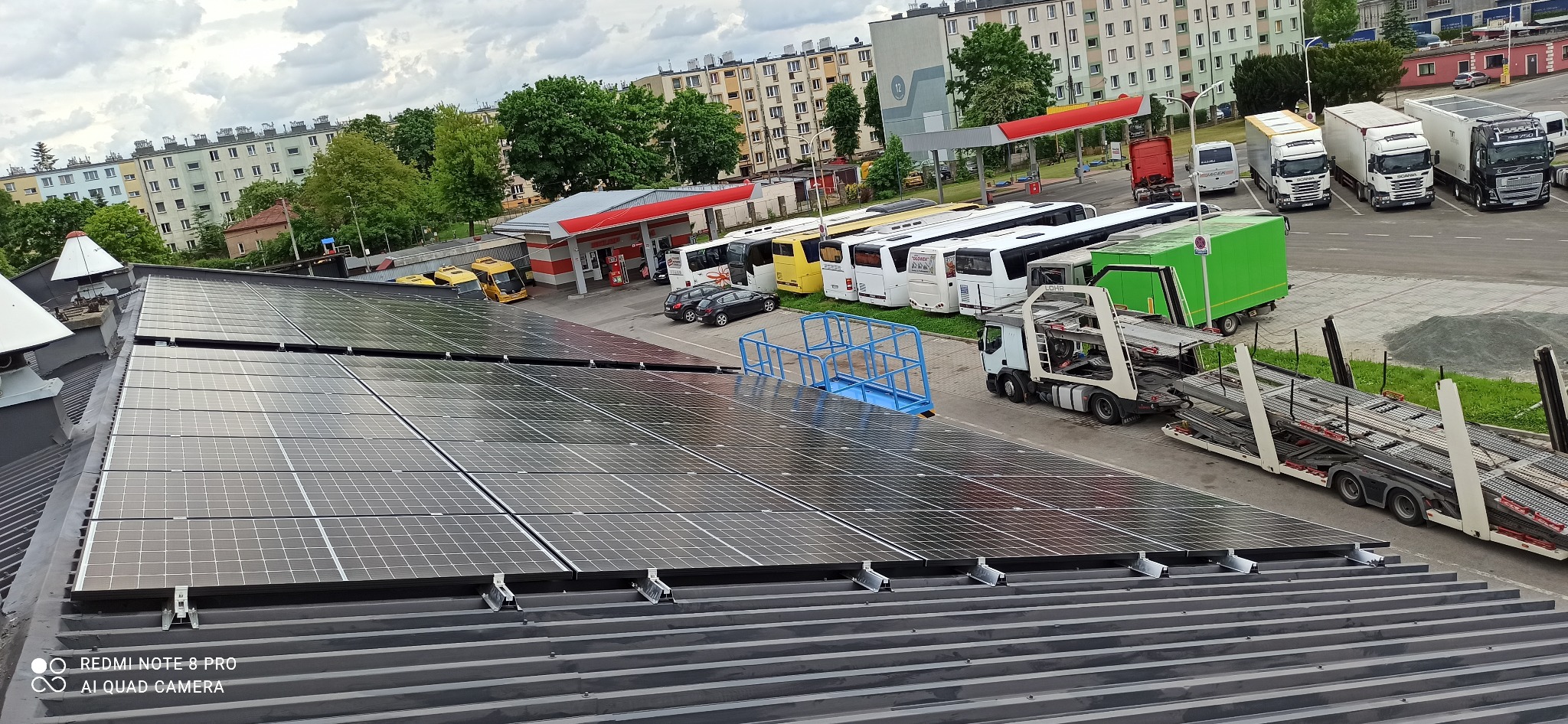 Panel słoneczny na dachu budynku, z widokiem na parking z autobusami i ciężarówkami w tle. Widoczne instalacje i mocowania paneli. Pochmurne niebo.