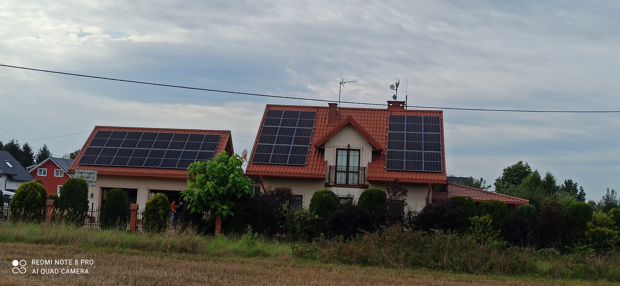 Dom jednorodzinny z czerwoną dachówką, na którym zamontowano panele słoneczne. Widoczne krzewy i drzewa w ogrodzie. Pochmurne niebo.