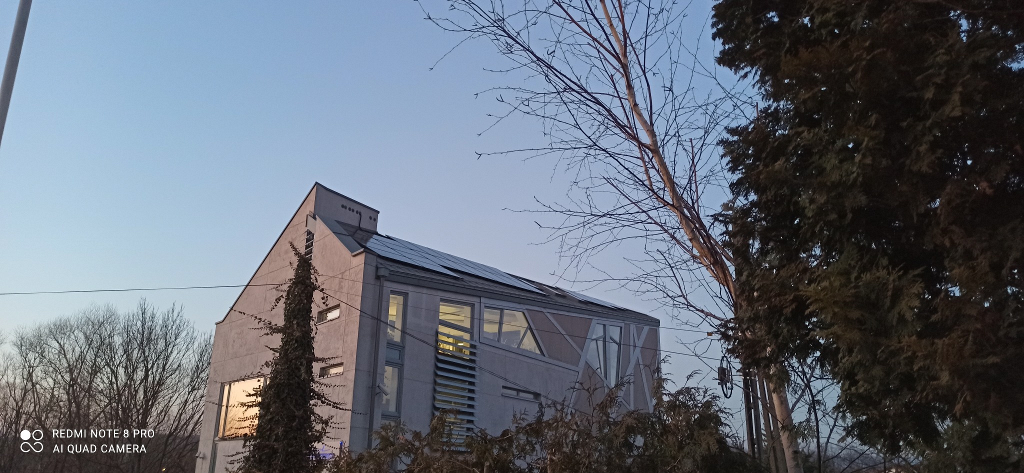 Dom z panelami słonecznymi na dachu, widziany z dołu, w tle błękitne niebo. W oknach odbija się zachodzące słońce, a z przodu rosną drzewa.