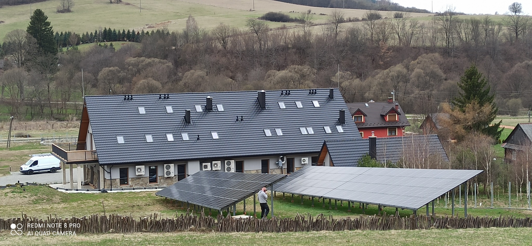 Instalacja paneli słonecznych na stelażach przed budynkiem z dachem pokrytym dachówką, w tle pagórkowaty krajobraz. Osoba przy panelach.