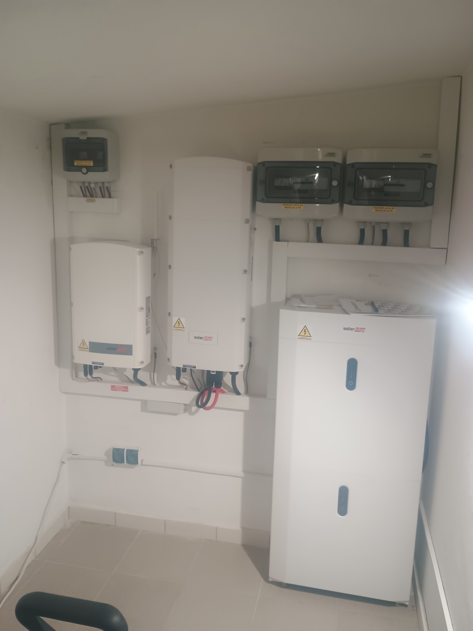 Białe urządzenia i rozdzielnice SolarEdge w pomieszczeniu, prawdopodobnie magazyn energii i inwertery. Widoczne przewody i ostrzeżenia o napięciu.