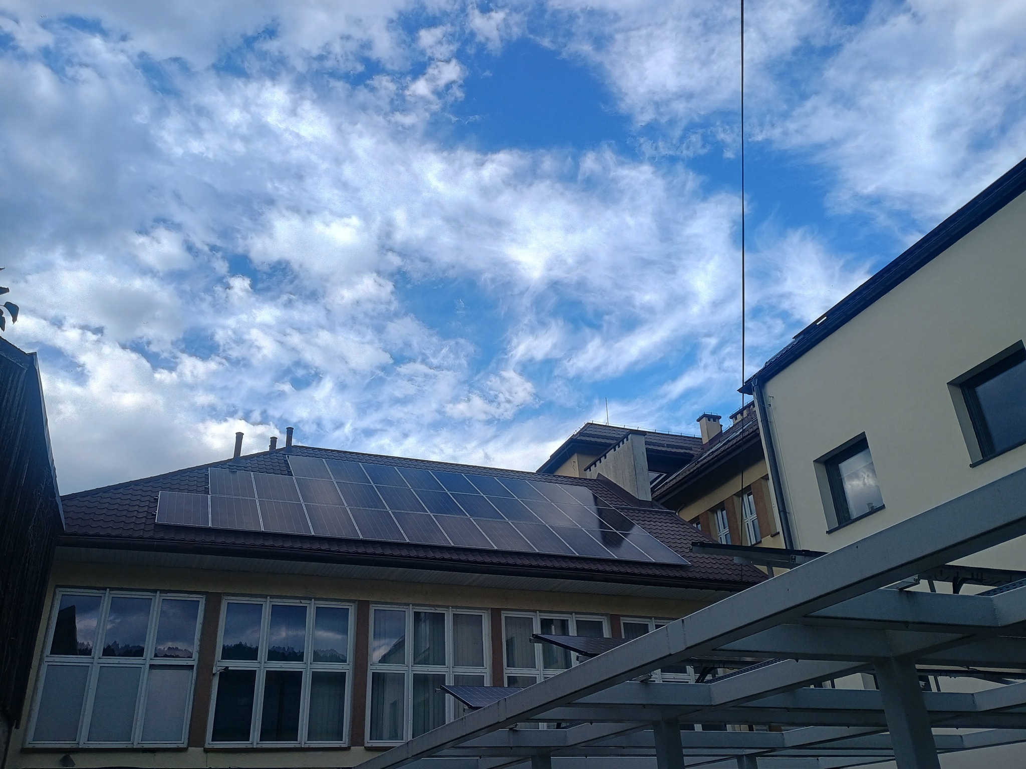 Panele słoneczne na dachu budynku z brązową dachówką, widok z dołu, błękitne niebo z chmurami w tle, metalowa konstrukcja w dolnej części kadru.