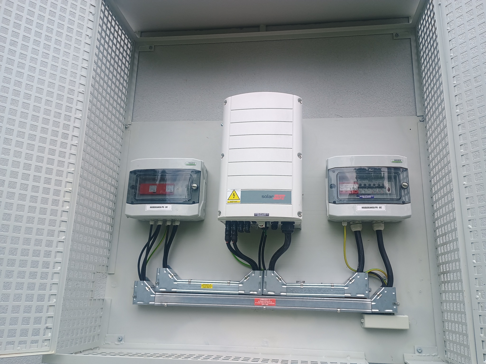 Kompletna instalacja fotowoltaiczna w obudowie: falownik SolarEdge pomiędzy dwoma rozdzielnicami PV-DC i PV-AC, kable w peszlach, szyna montażowa z ostrzeżeniami.