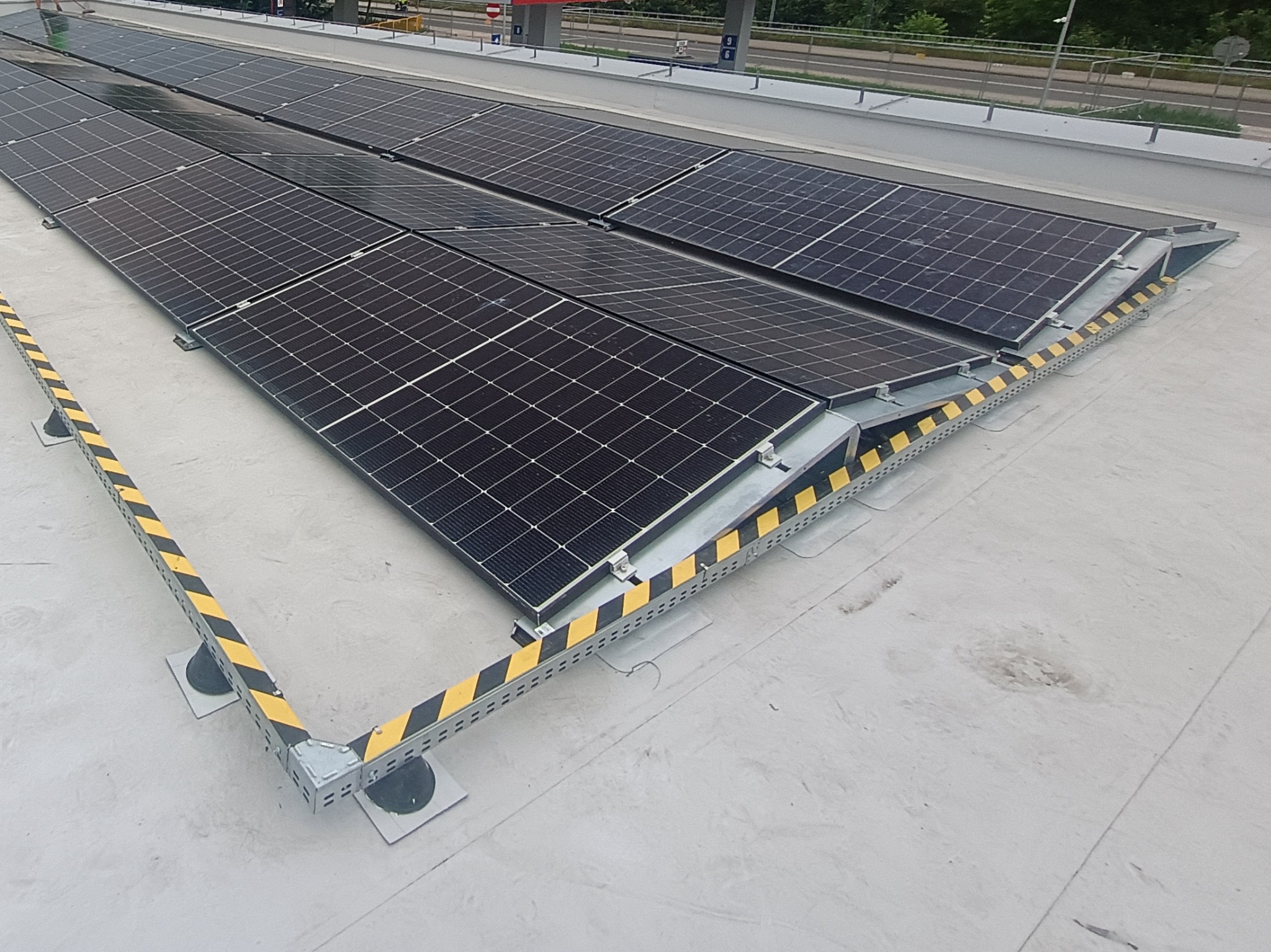 Rząd paneli słonecznych na dachu, zabezpieczony żółto-czarną barierą. Widok z góry, z widoczną konstrukcją montażową paneli i elementami zabezpieczeń.