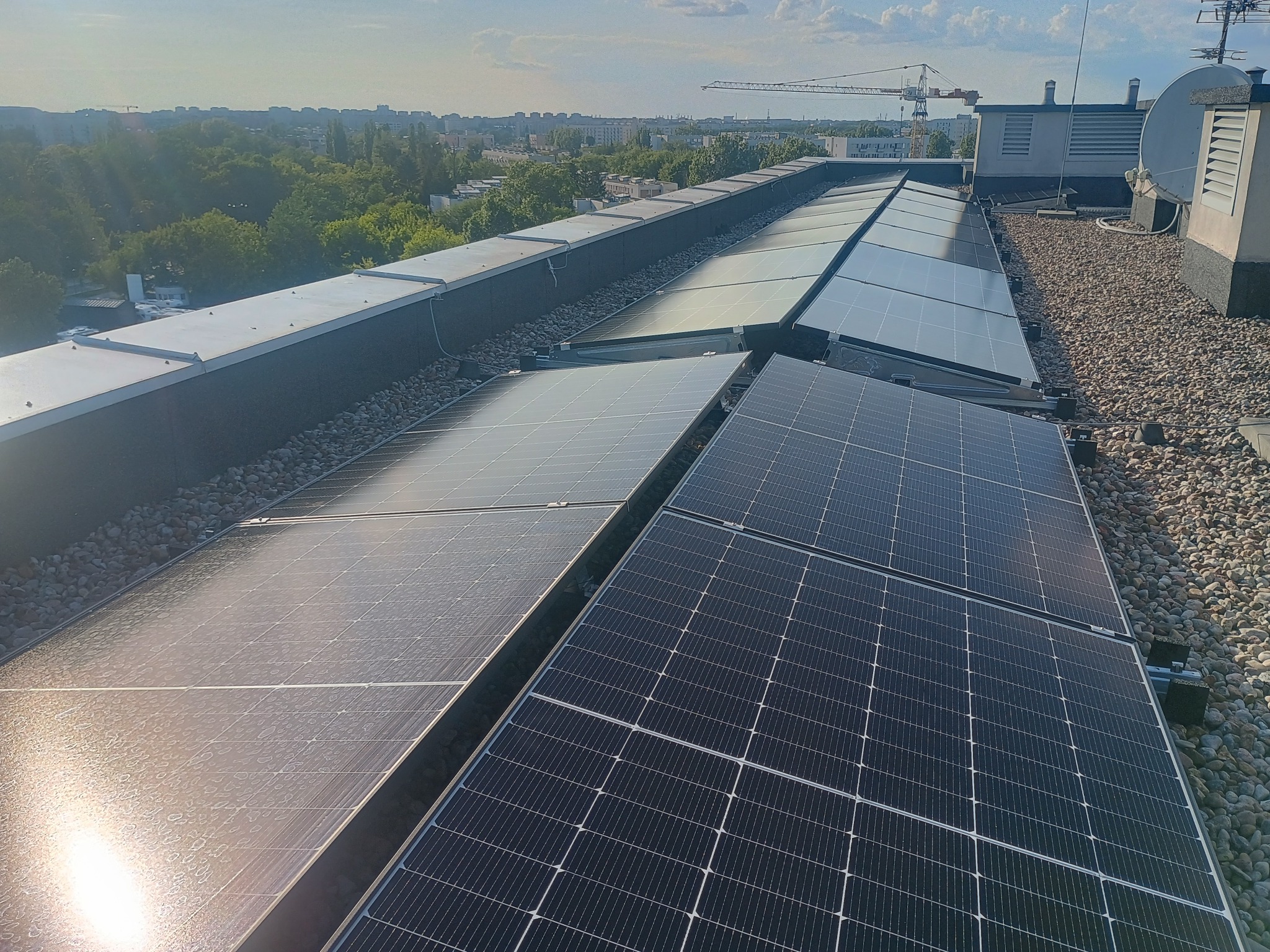 Rząd paneli słonecznych na płaskim dachu budynku, otoczonych drobnymi kamieniami. W tle widoczny krajobraz miejski i niebo.