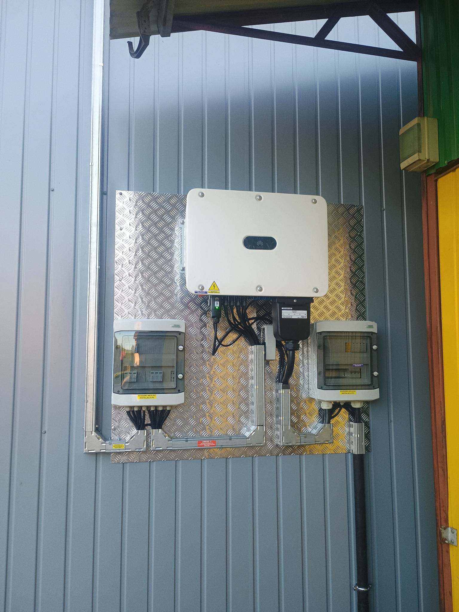 Instalacja fotowoltaiczna: inwerter i rozdzielnice DC/AC zamontowane na ścianie z blachy trapezowej, widoczne kable i oznaczenia.