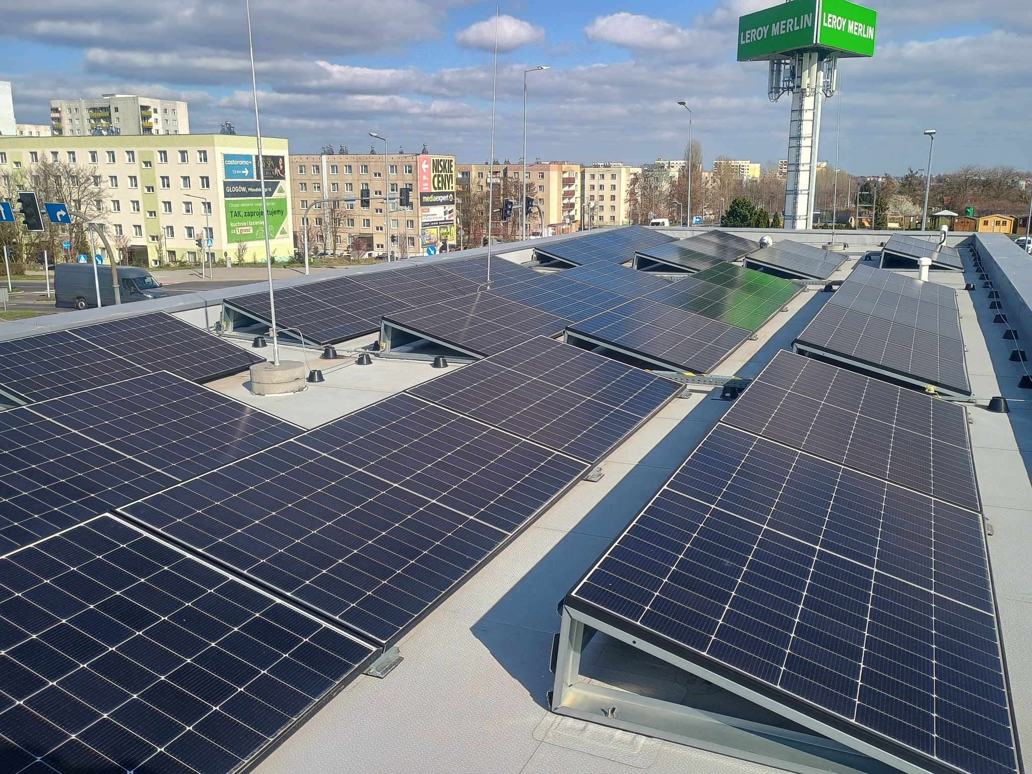 Instalacja paneli słonecznych na dachu budynku handlowego Leroy Merlin, widok z góry, słoneczny dzień. Panele ułożone rzędami, w tle budynki mieszkalne i niebieskie niebo.