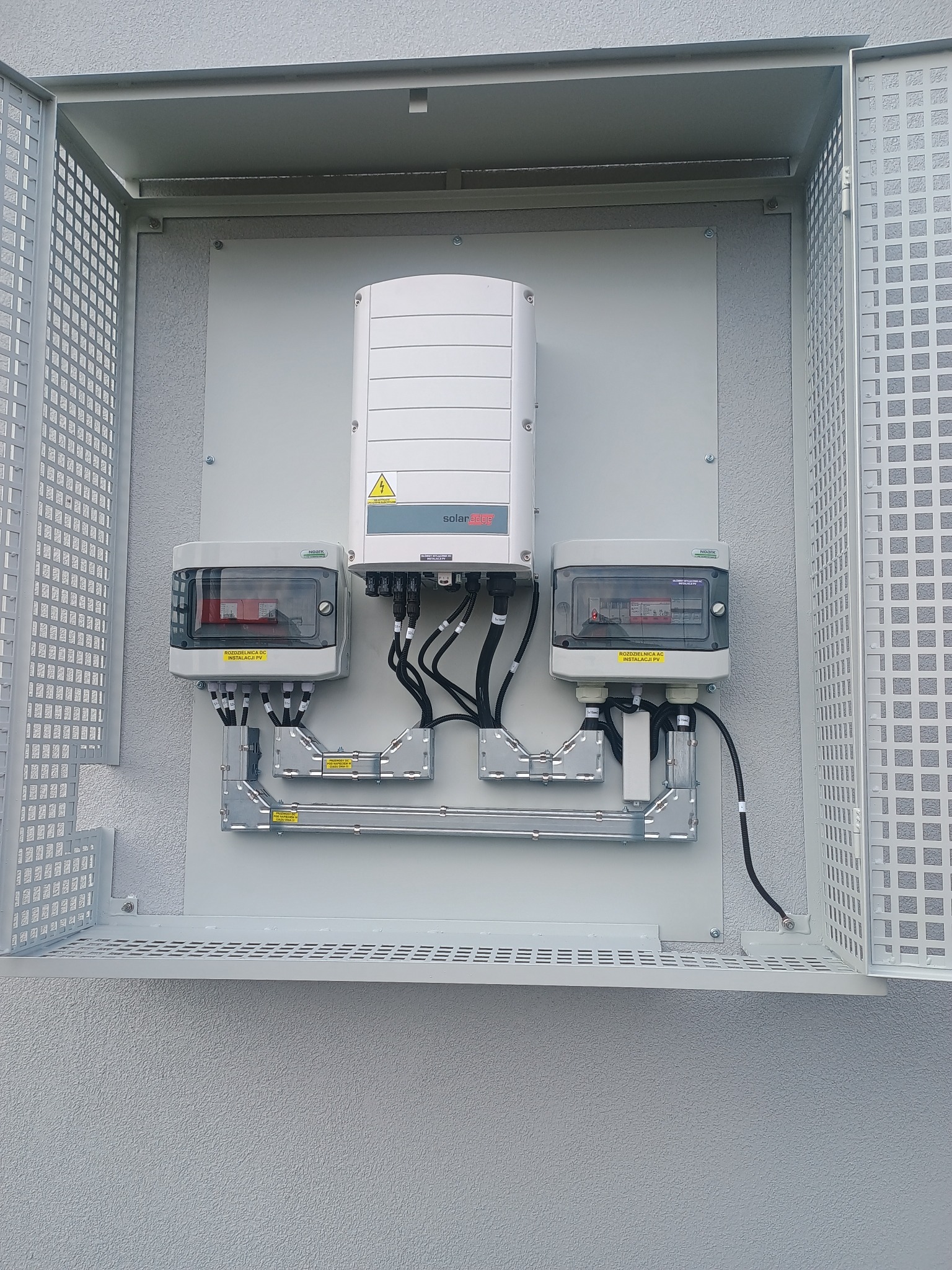 Kompletna instalacja fotowoltaiczna w szarej obudowie z siatki ochronnej, z widocznym inwerterem i rozdzielnicami AC/DC, zamontowana na elewacji budynku.