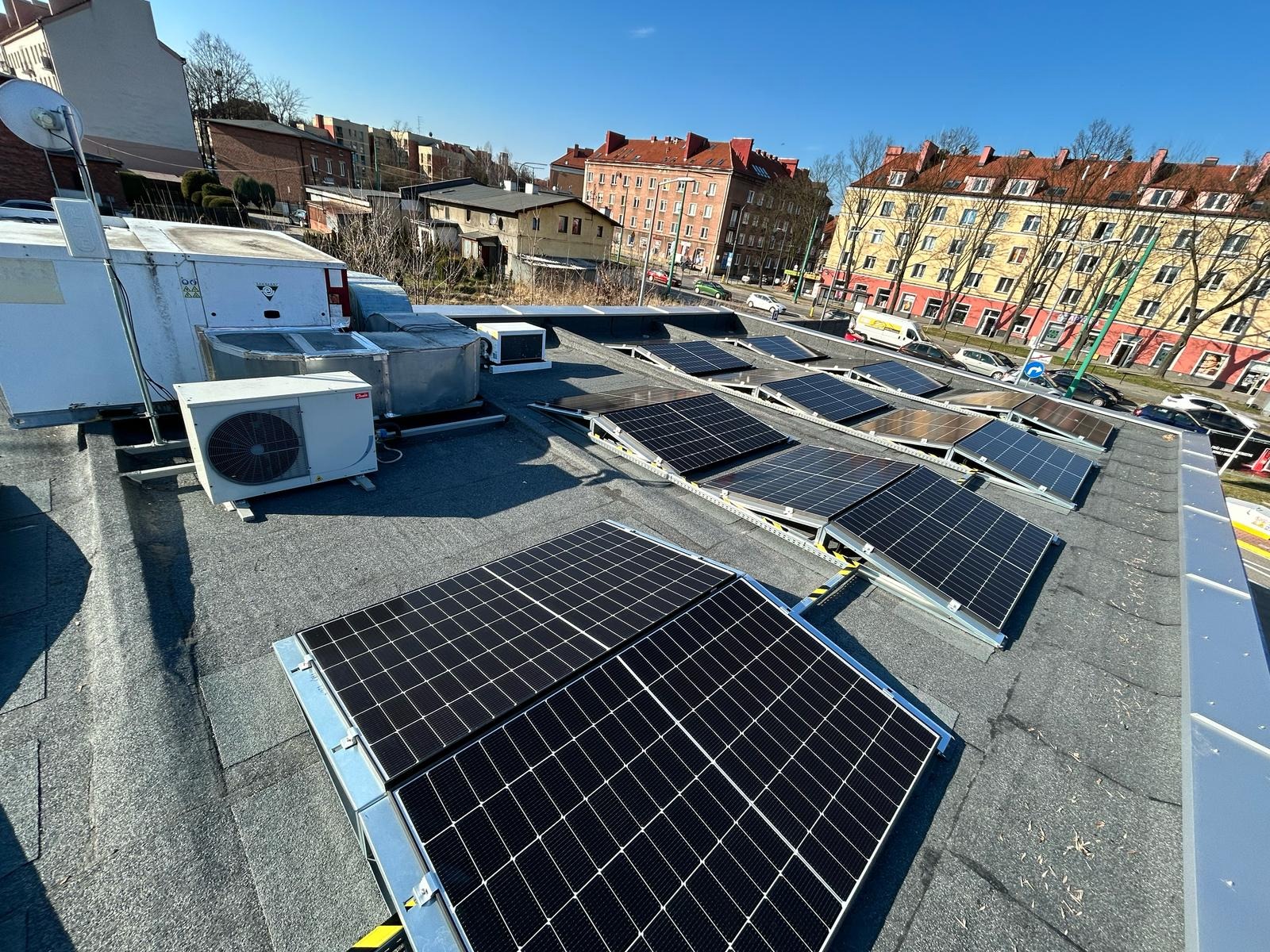 Instalacja paneli fotowoltaicznych na dachu budynku miejskiego z widocznymi jednostkami klimatyzacyjnymi i zabudową miejską w tle, w słoneczny dzień.