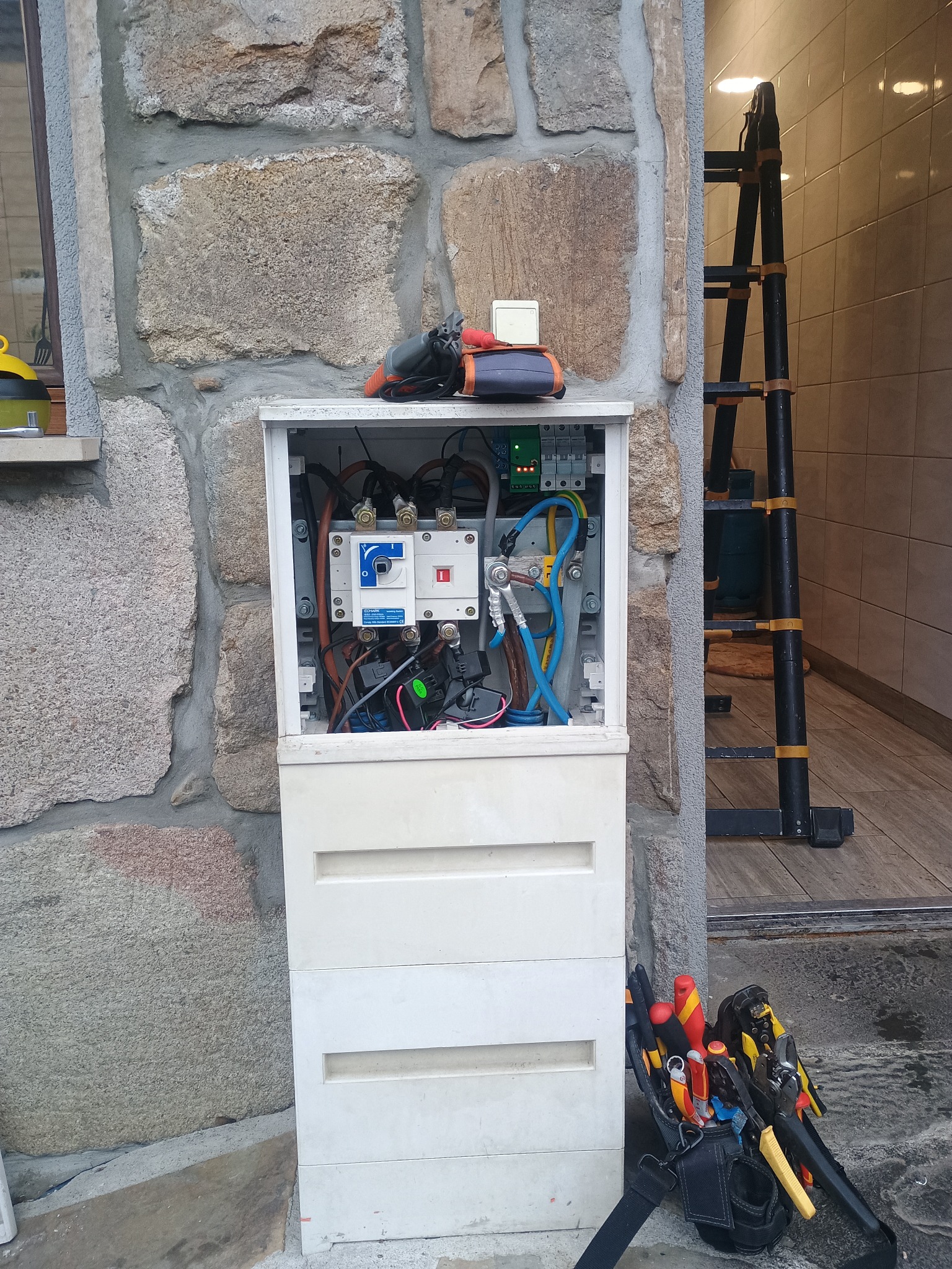 Otwarta skrzynka elektryczna na kamiennej ścianie budynku, widoczne kable, bezpieczniki i narzędzia elektryczne. Obok drabina i torba z narzędziami.