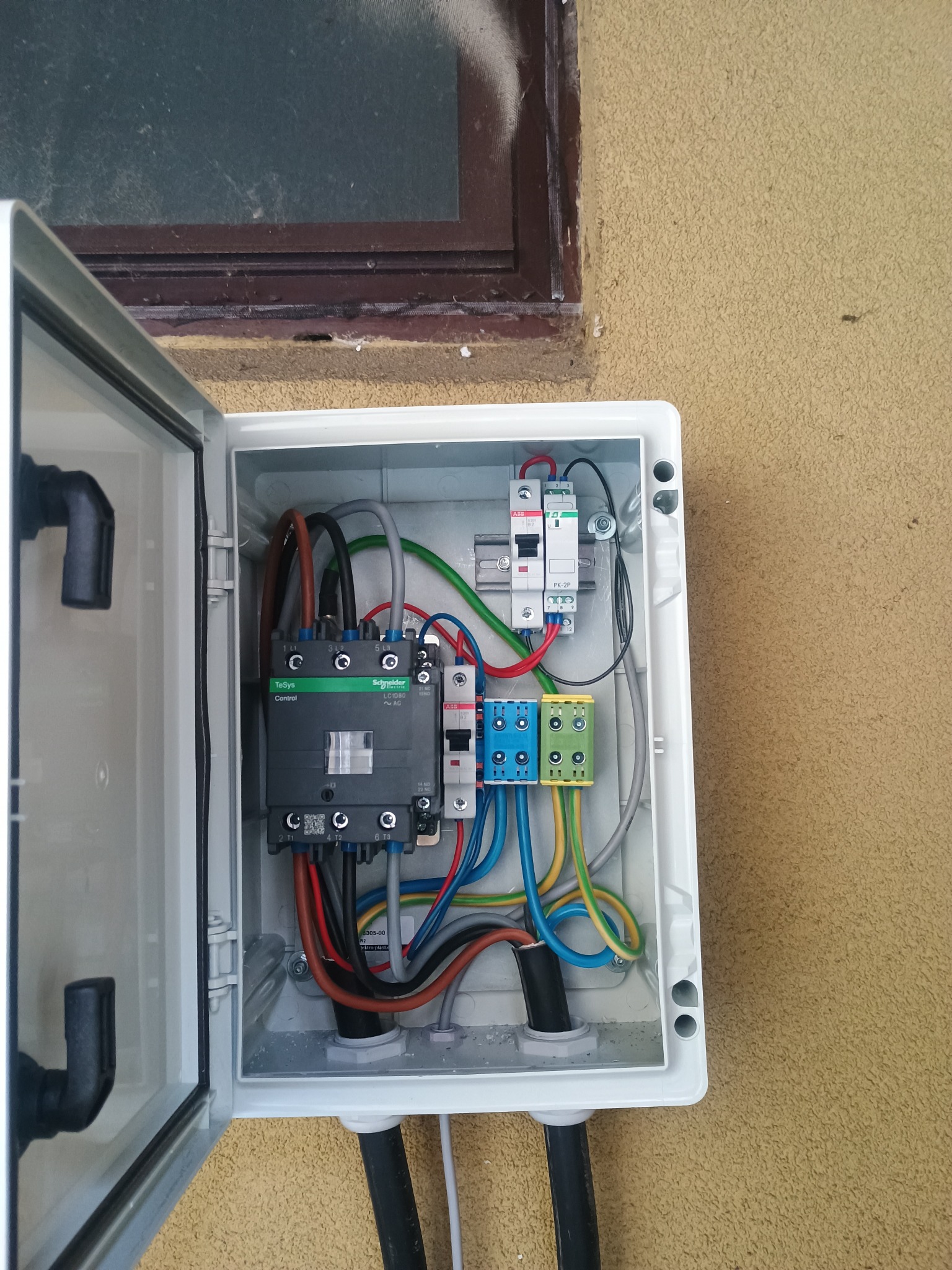 Otwarta, biała skrzynka elektryczna z widocznymi bezpiecznikami, stycznikiem Schneider Electric i kolorowymi przewodami. Skrzynka zamontowana na elewacji budynku obok okna.