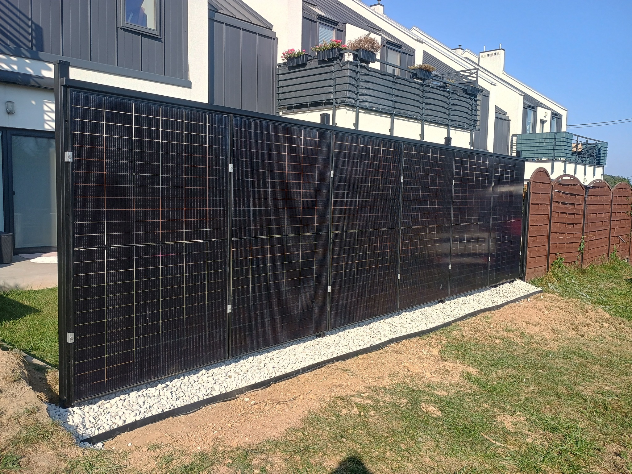 Panele fotowoltaiczne zamontowane jako ogrodzenie posesji. Widoczne czarne ramy, jasne kamienie u podstawy i fragmenty budynków w tle, w słoneczny dzień.