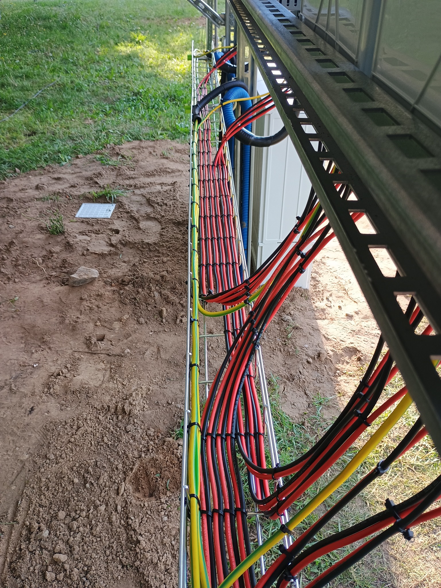 Uporządkowane kable w metalowej korycie kablowej, równolegle ułożone, zabezpieczone opaskami, widoczne kolory: czerwony, czarny, żółto-zielony. Tło: trawa, ziemia.