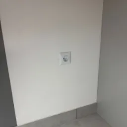 Białe gniazdko elektryczne typu francuskiego na świeżo pomalowanej, gładkiej, białej ścianie nad szarą listwą przypodłogową i płytkami podłogowymi.