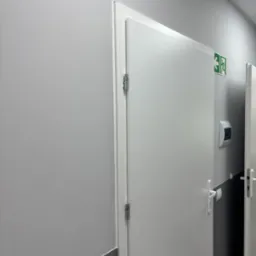 Białe drzwi z metalową klamką i zawiasami w jasnym pomieszczeniu. Nad drzwiami widoczny znak ewakuacyjny. Ściana pomalowana na szaro.