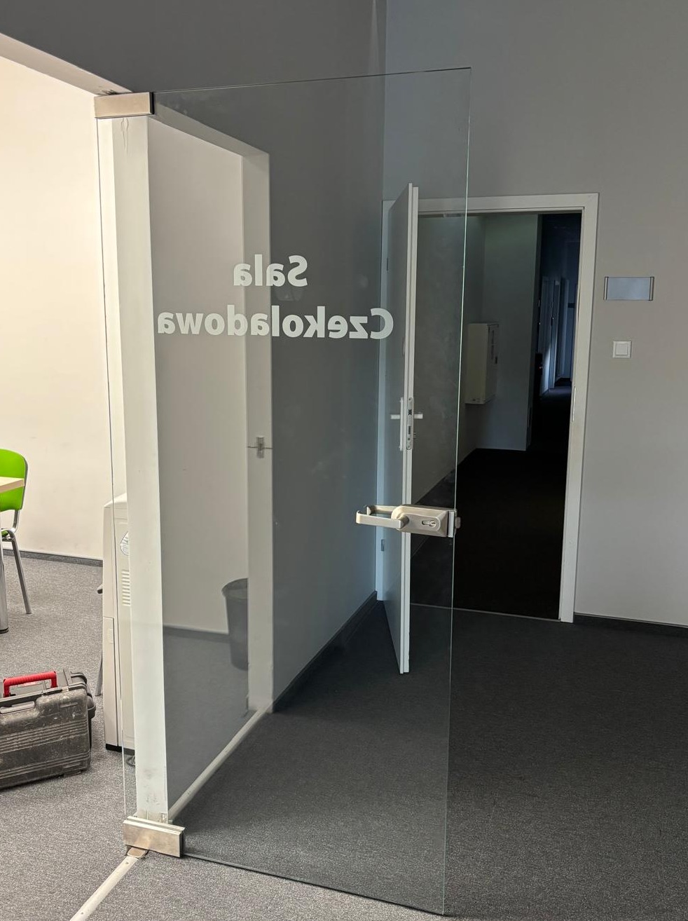Szklane drzwi z napisem w biurze we Wrocławiu. Widoczny metalowy zawias i klamka. W tle inne pomieszczenia i drzwi. Na podłodze czarna wykładzina.