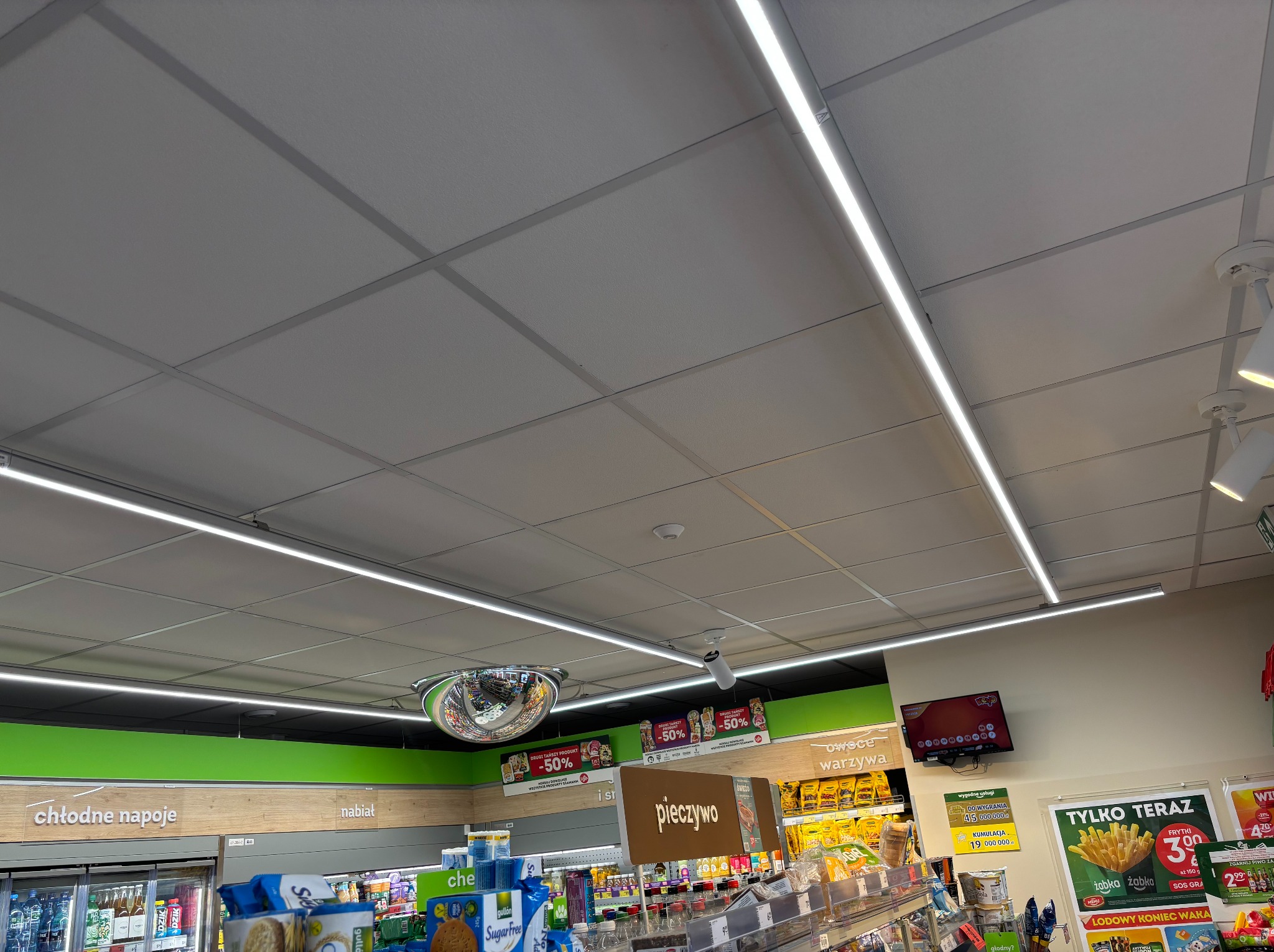 Sufit podwieszany z rastrem i oświetleniem LED w supermarkecie. Widoczne elementy instalacji, takie jak czujniki i kamery. Perspektywa od dołu, oświetlenie jasne.
