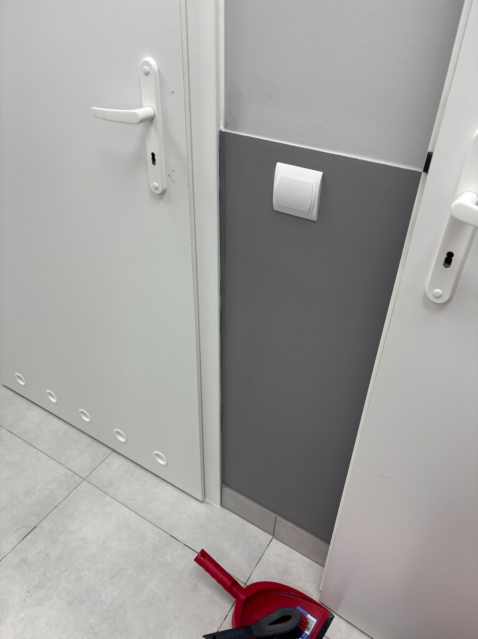 Białe drzwi z klamką i listwą przypodłogową, obok ściana z włącznikiem. Na podłodze leży szufelka z zmiotką. Widok z dołu, po remoncie.