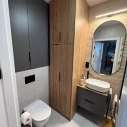 Nowoczesna łazienka po remoncie: szare szafki, drewnopodobna zabudowa, owalne lustro z podświetleniem, biała ceramika i płytki w odcieniach szarości.