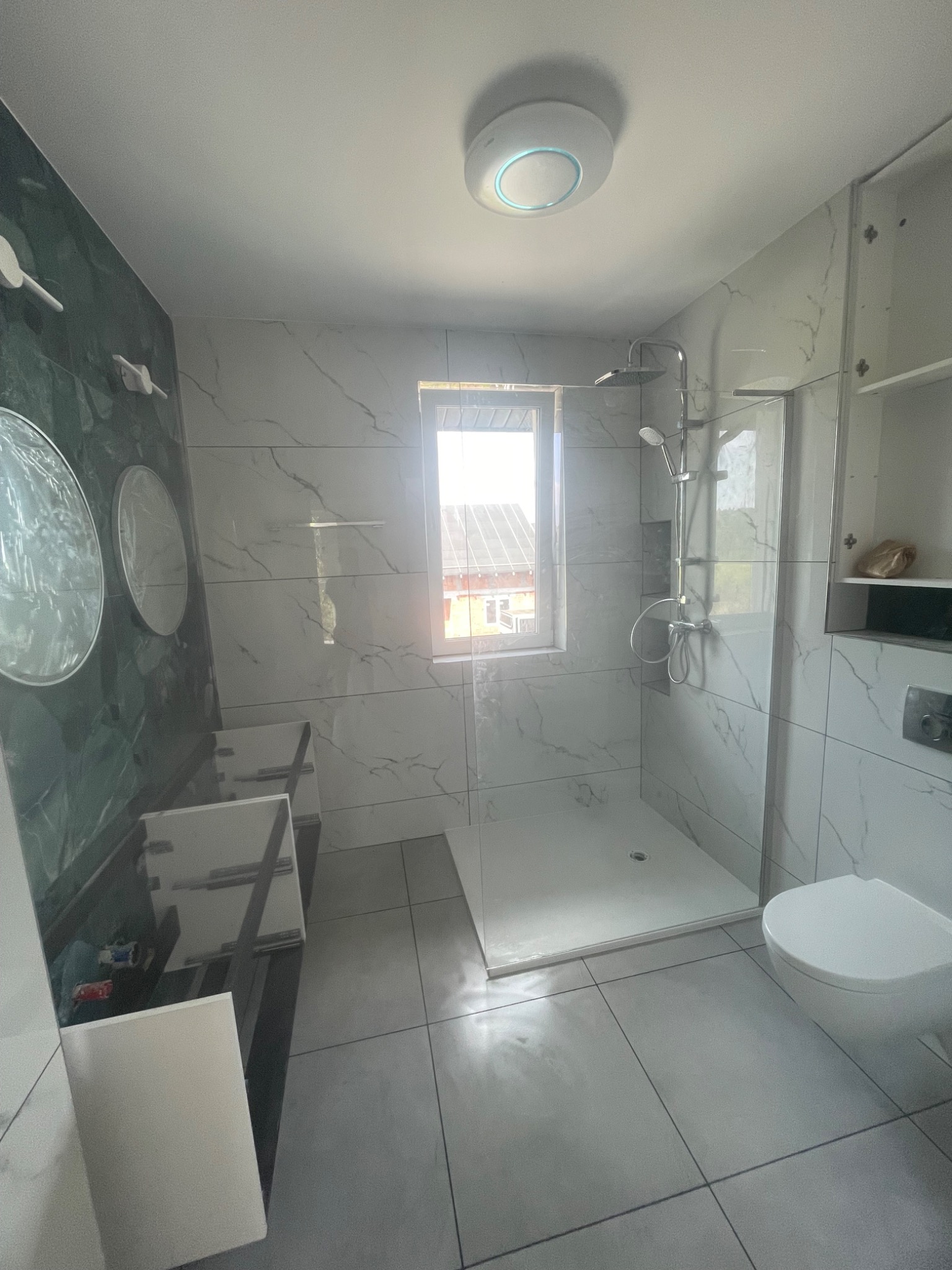 Nowoczesna łazienka z kabiną prysznicową, marmurowymi płytkami, umywalką z szafką i okrągłymi lustrami. Widoczne detale wykończenia i oświetlenie sufitowe.