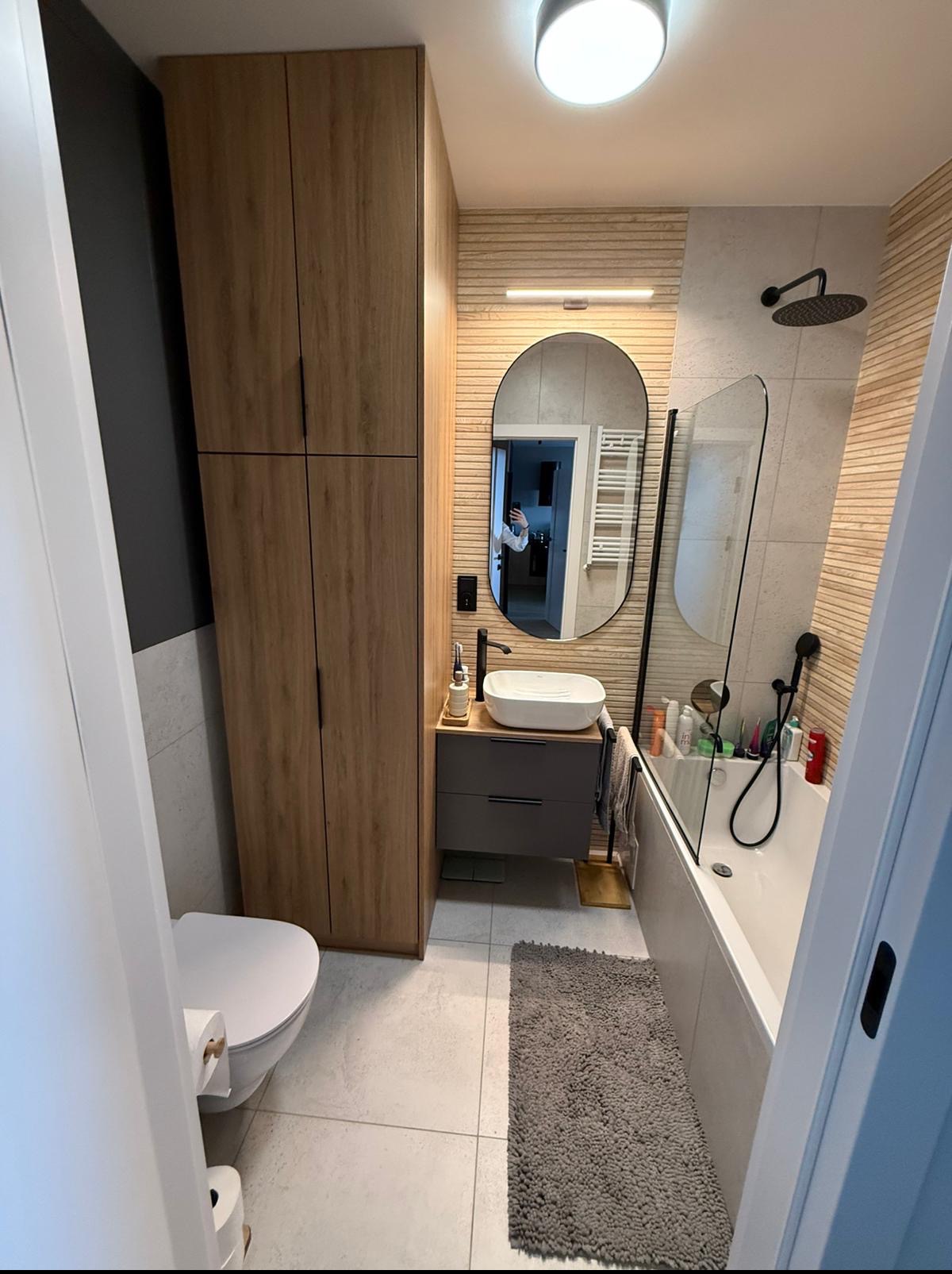 Nowoczesna łazienka z drewnianą szafką, owalnym lustrem i beżowymi płytkami imitującymi drewno. Minimalistyczna umywalka nablatowa i czarna armatura dodają elegancji.