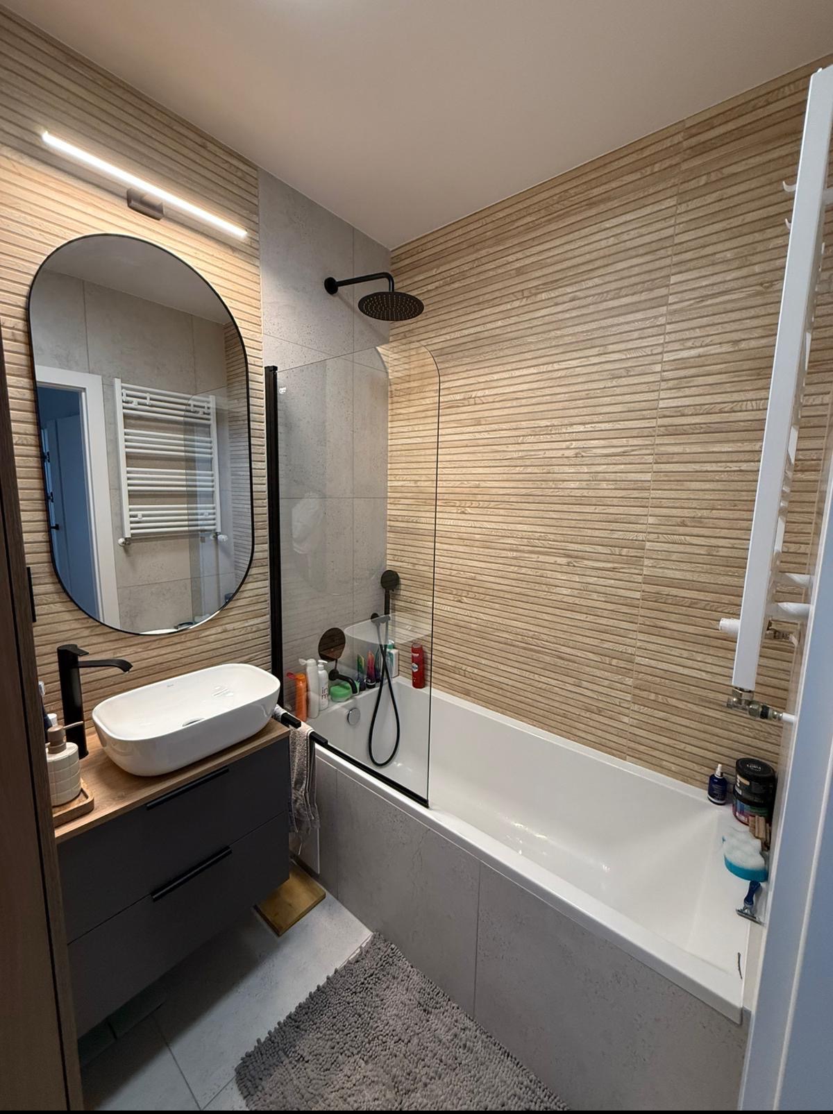 Ukończona łazienka: beżowe płytki imitujące drewno na ścianie, owalne lustro z podświetleniem, biała wanna z czarną armaturą i szklanym panelem.