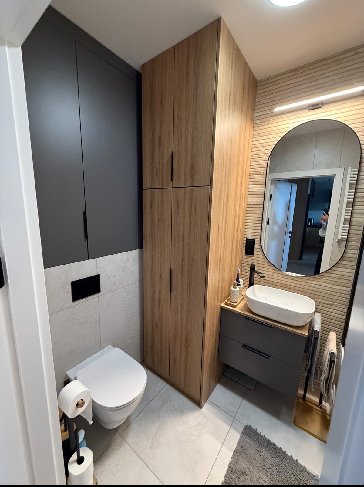 Nowoczesna łazienka po remoncie: szare szafki, drewnopodobna zabudowa, owalne lustro z podświetleniem, biała ceramika i płytki w odcieniach szarości.