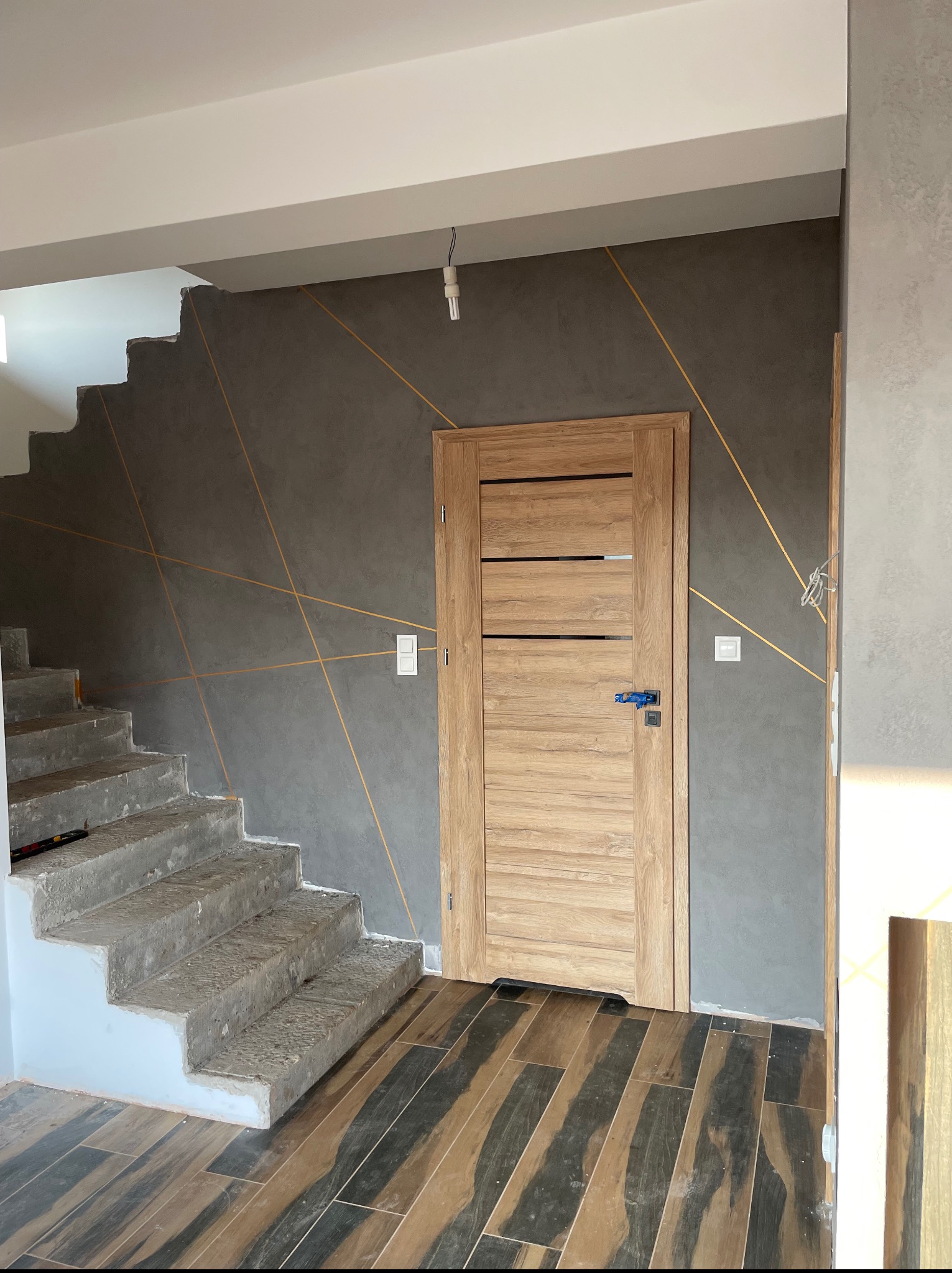 Szare ściany z geometrycznym złotym wzorem, drewniane drzwi i schody w trakcie prac. Podłoga wyłożona płytkami imitującymi drewno. Widoczne gniazdka i surowy stan schodów.