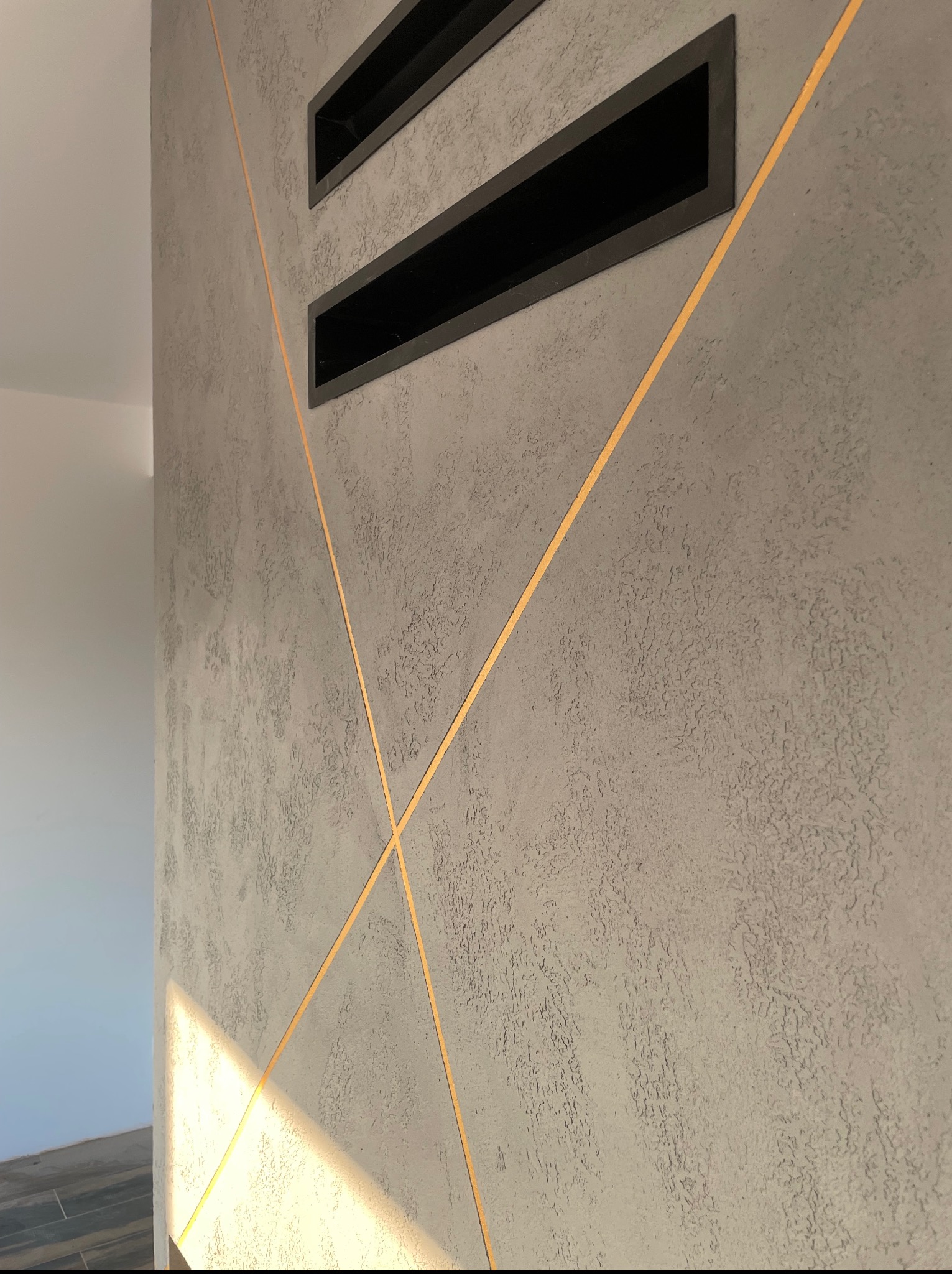 Dekoracyjna ściana z szarego tynku strukturalnego, zdobiona złotymi liniami krzyżującymi się i czarnymi wnękami, oświetlona naturalnym światłem.