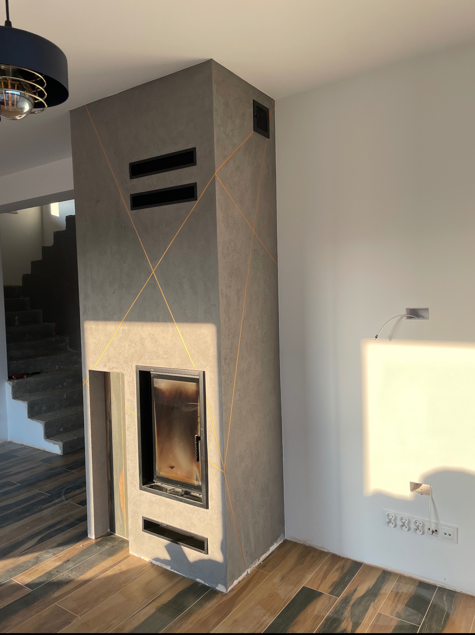 Wykończony kominek z szarą strukturą i złotymi liniami na tle białej ściany. Widoczne gniazdka elektryczne i drewnopodobna podłoga. Częściowo widoczne schody.