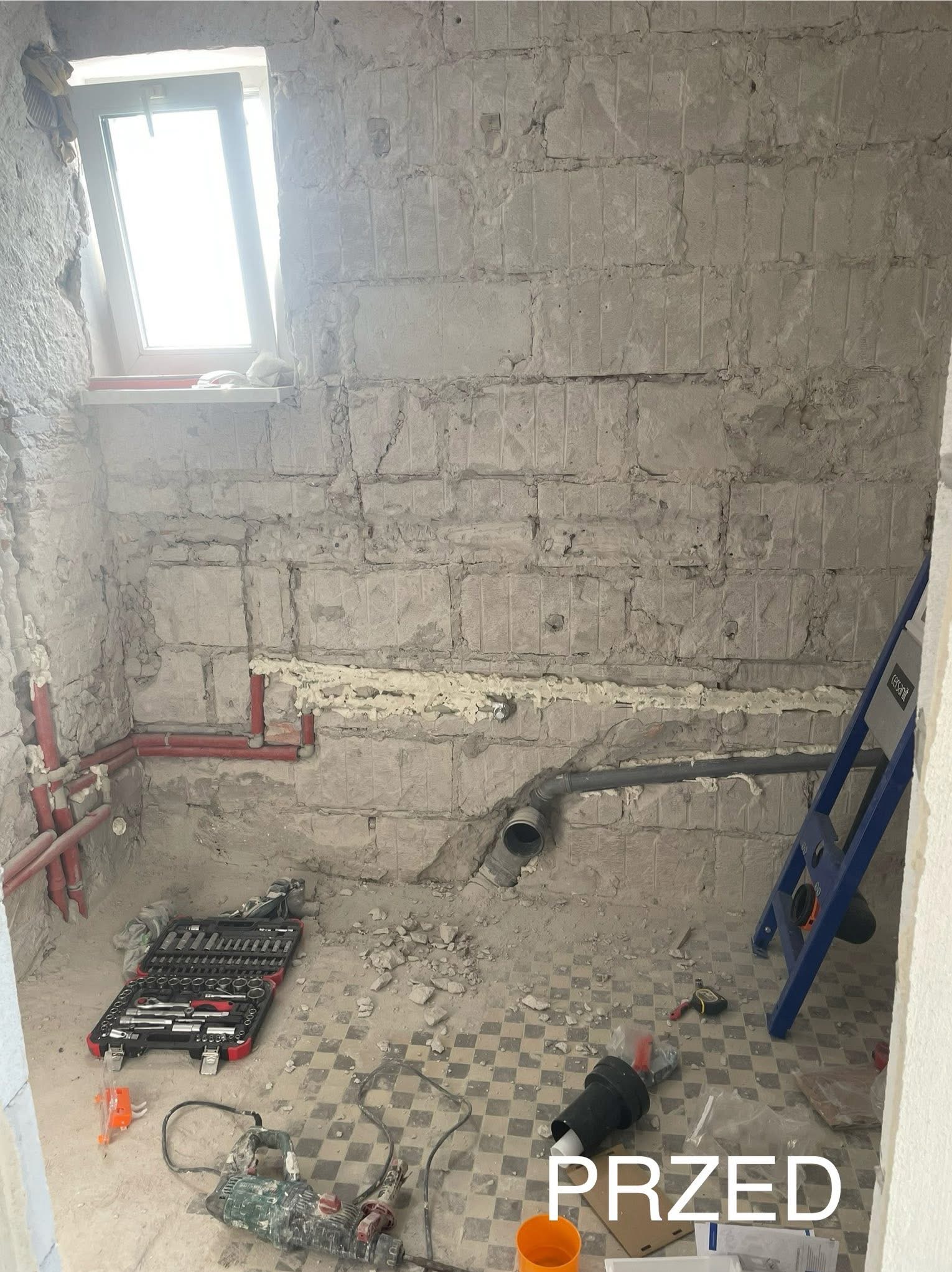Łazienka w trakcie remontu: surowe ściany z widocznymi instalacjami, narzędzia i drabina. Stan 'przed' z widocznym oknem i starą podłogą w kratkę.