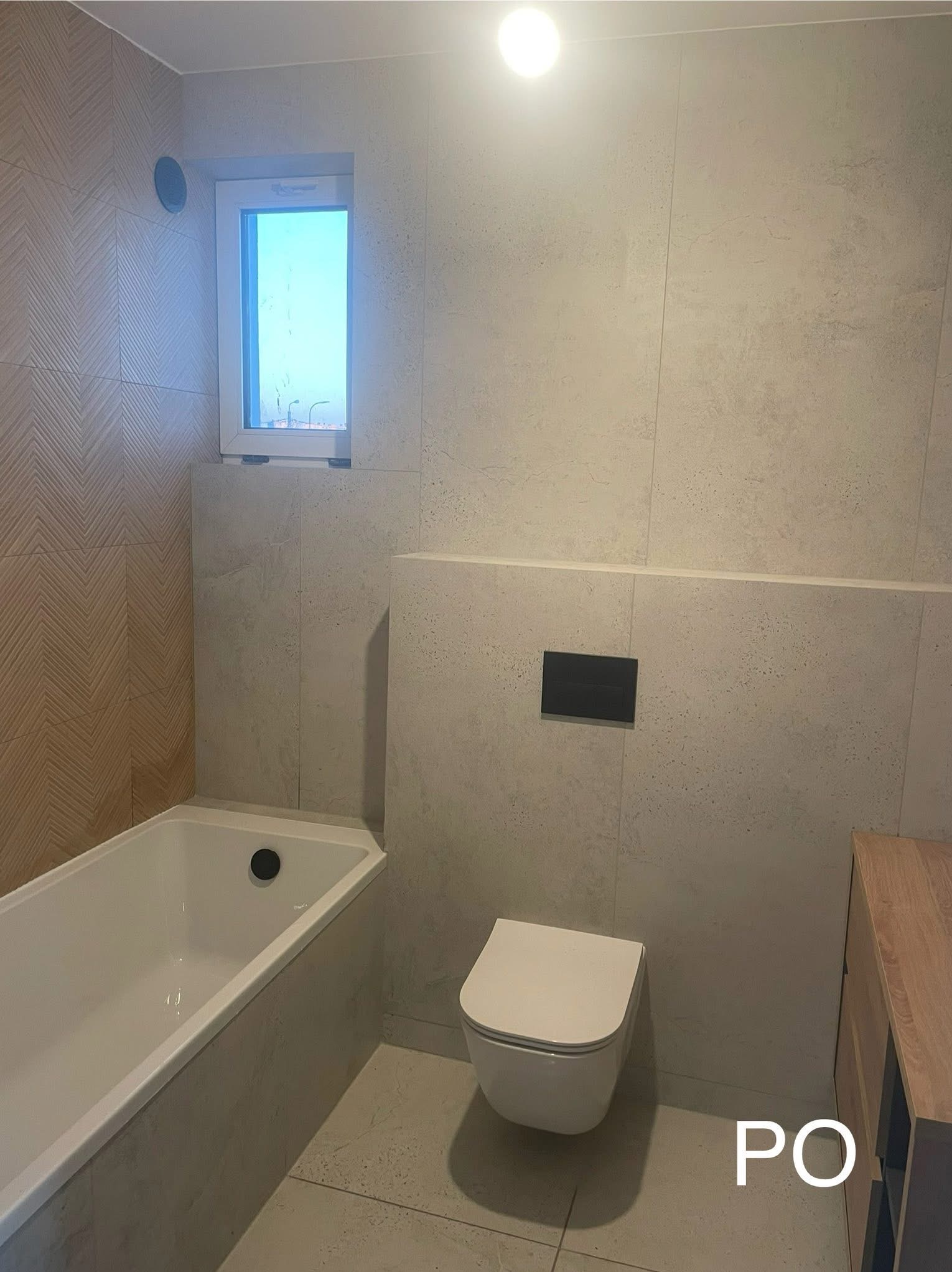 Wykończona łazienka z wanną, toaletą podwieszaną i szafką z drewnianym frontem. Na ścianach jasne płytki, w tym jedna ściana z wzorem w jodełkę. Małe okno z widokiem na zewnątrz.