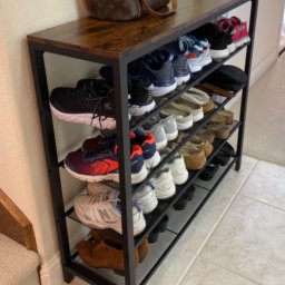 Roman Ko - Metalowy regał na buty z drewnianym blatem, wypełniony różnorodnym obuwiem, obok schodów i torby. Minimalistyczny design, jasne płytki podłogowe.